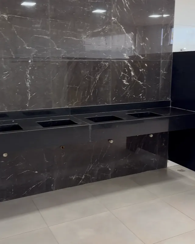 Banheiro do complexo: bancada de granito preto com quatro cubas e parede de porcelanato marmorizado.