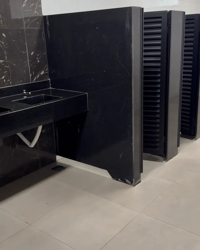 Banheiro do complexo: bancada com cuba e cabines com portas pretas.