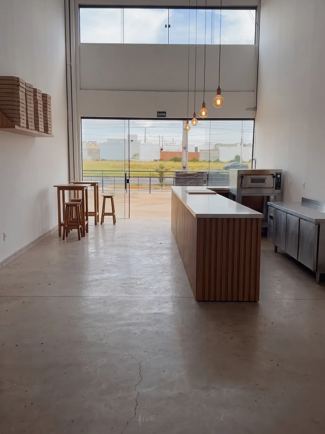Imagem conceitual de ocupação: a mesma loja com balcão de madeira ripada, forno de pizza e bancada de inox, mesas altas junto à vitrine e caixas de pizza numa prateleira.